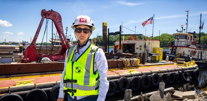 Elizabeth Wilkinson wears a bright yellow safety vest and helmet stands in front of a barge at a dock on a sunny day, with a tugboat and American flag visible in the background.
