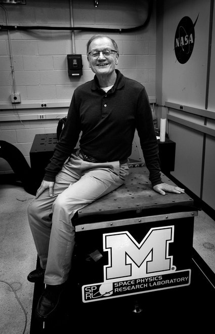 Smiling researcher sits on a blue laboratory platform marked with a large University of Michigan “M” logo and NASA insignia inside a research facility.