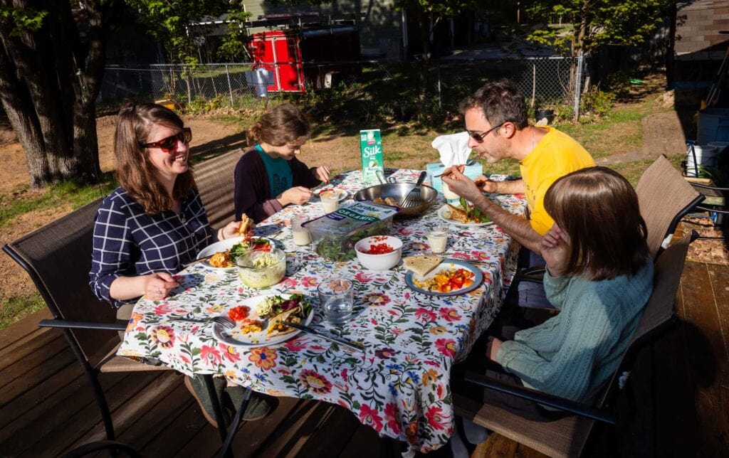 Four people share a meal outdoors around a table, gathered in a wooded setting.