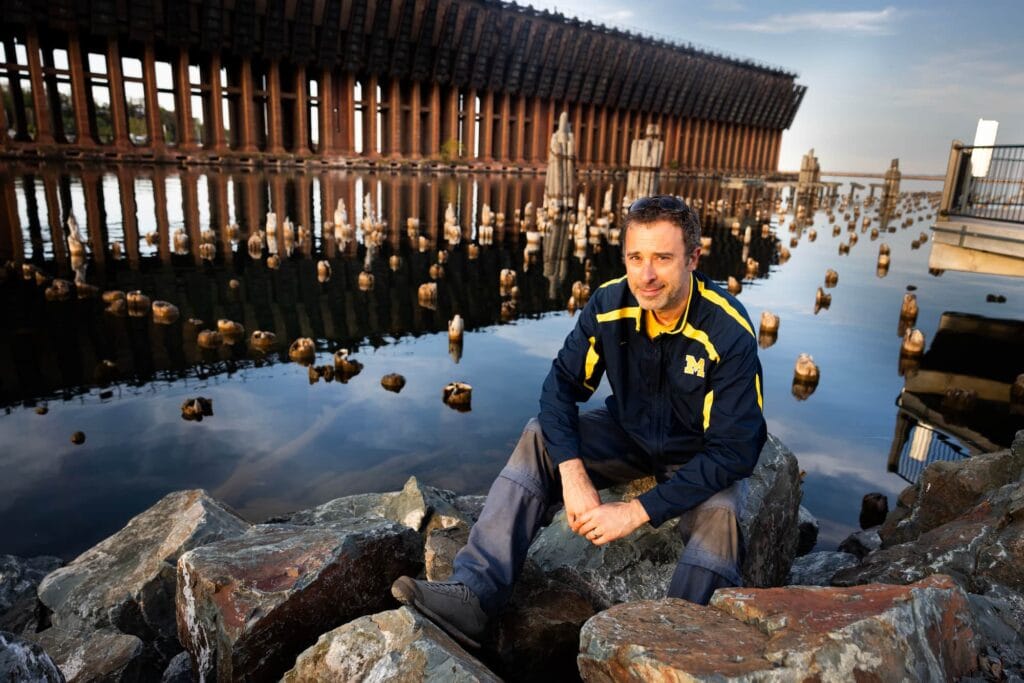 Pete Cook sits on rocks along a waterfront with a large industrial ore dock structure in the background.