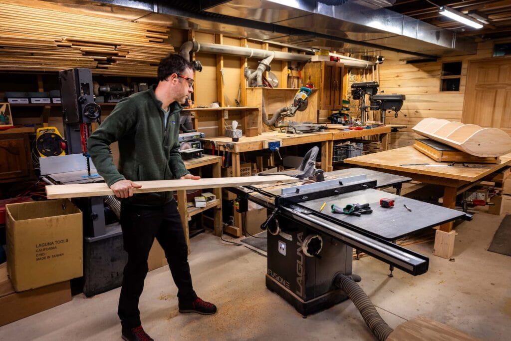 Pete Cook works in his woodworking shop, operating a table saw surrounded by stacks of lumber and tools.