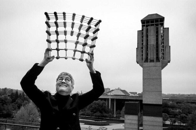 Elizabeth A. Holm holds a ball-and-stick model of the atomic structure of a material over her head. A tower stands in the background.