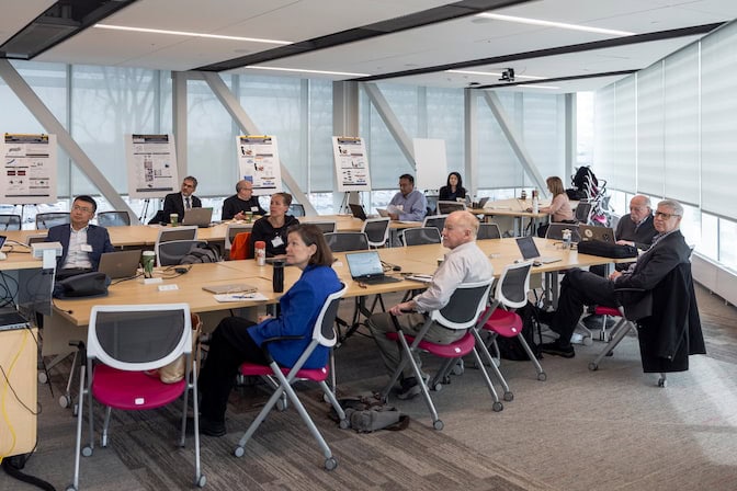 11 people sit at modular conference tables, facing a speaker out of frame, with posters along one wall.