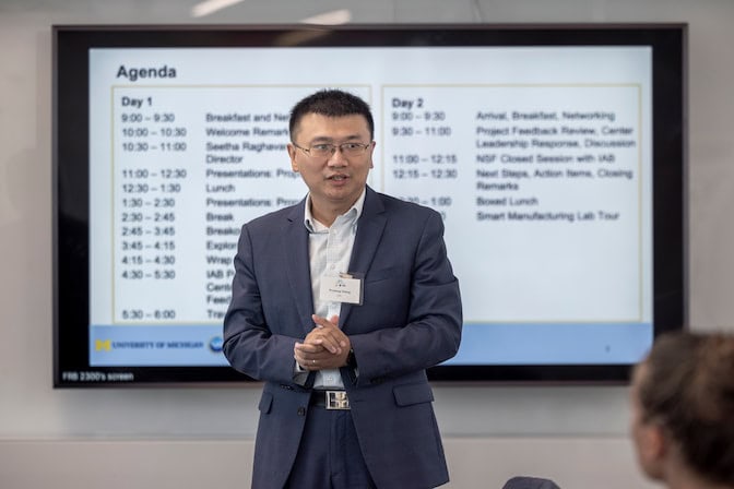 A man stands with hands clasped in front of the screen, which displays the Day 1 and Day 2 agenda.