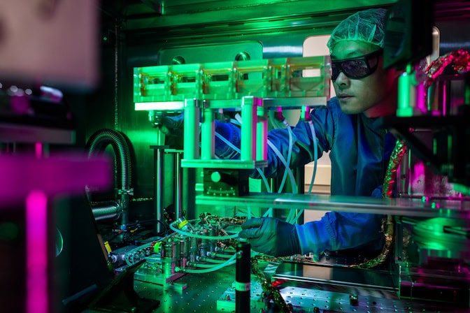 A man in laser safety goggles leans into a cabinet containing an optical table with tubes running between components.