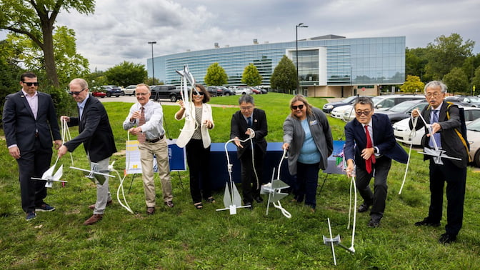 Eight adults participate in a groundbreaking ceremony outside a modern glass building, holding ropes attached to small ship anchors.