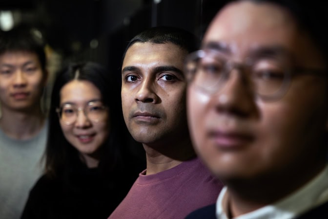 Four people stand in a line, looking cool as heck. The camera is tightly focused on one man's face. The other people are slightly out of focus.
