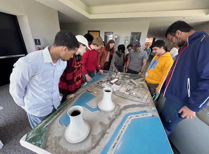 A group of students gathers around a detailed model of a nuclear power plant featuring two cooling towers.
