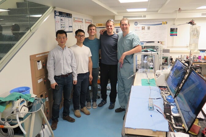 Five men stand in a lab near a box with some tubes and wires coming out of it.