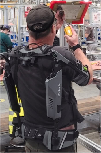  A man wearing a shoulder exoskeleton holds a drill in one hand pointing toward a metal piece positioned with the other hand on an angled sheet of metal at his eye level.
