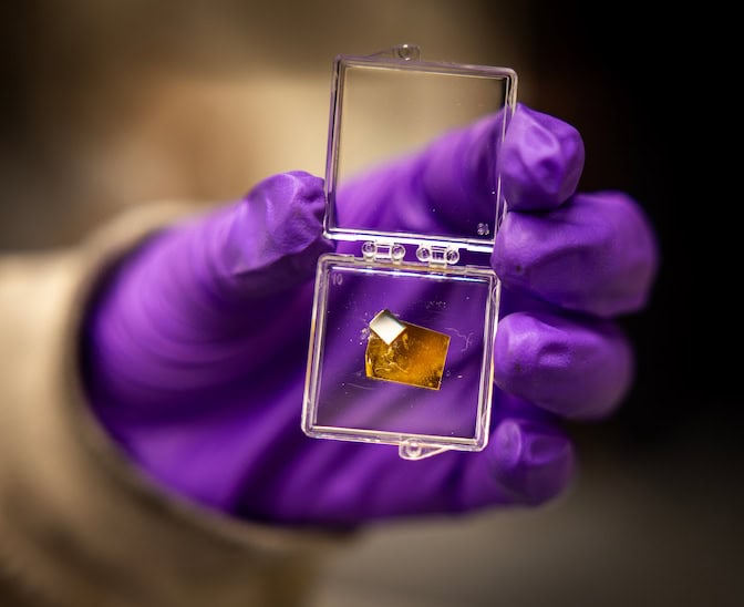 A hand clad in a bright purple glove holds a clear plastic case. The case's hinged lid is open, revealing a small square of metal inside.